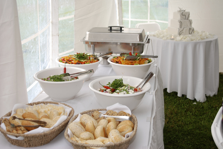 Buffet setup under a white tent: bowls of colorful salads, baskets of bread rolls, and a stainless chafing dish on a long table.